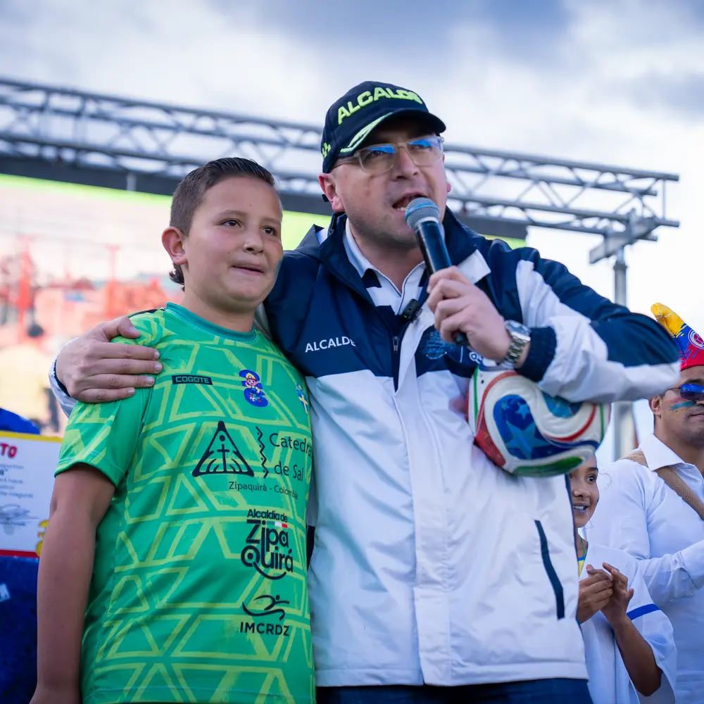 El alcalde de Zipaquirá habla por micrófono abrazando a un niño futbolista con la camiseta oficial patrocinada por la Catedral de Sal.
