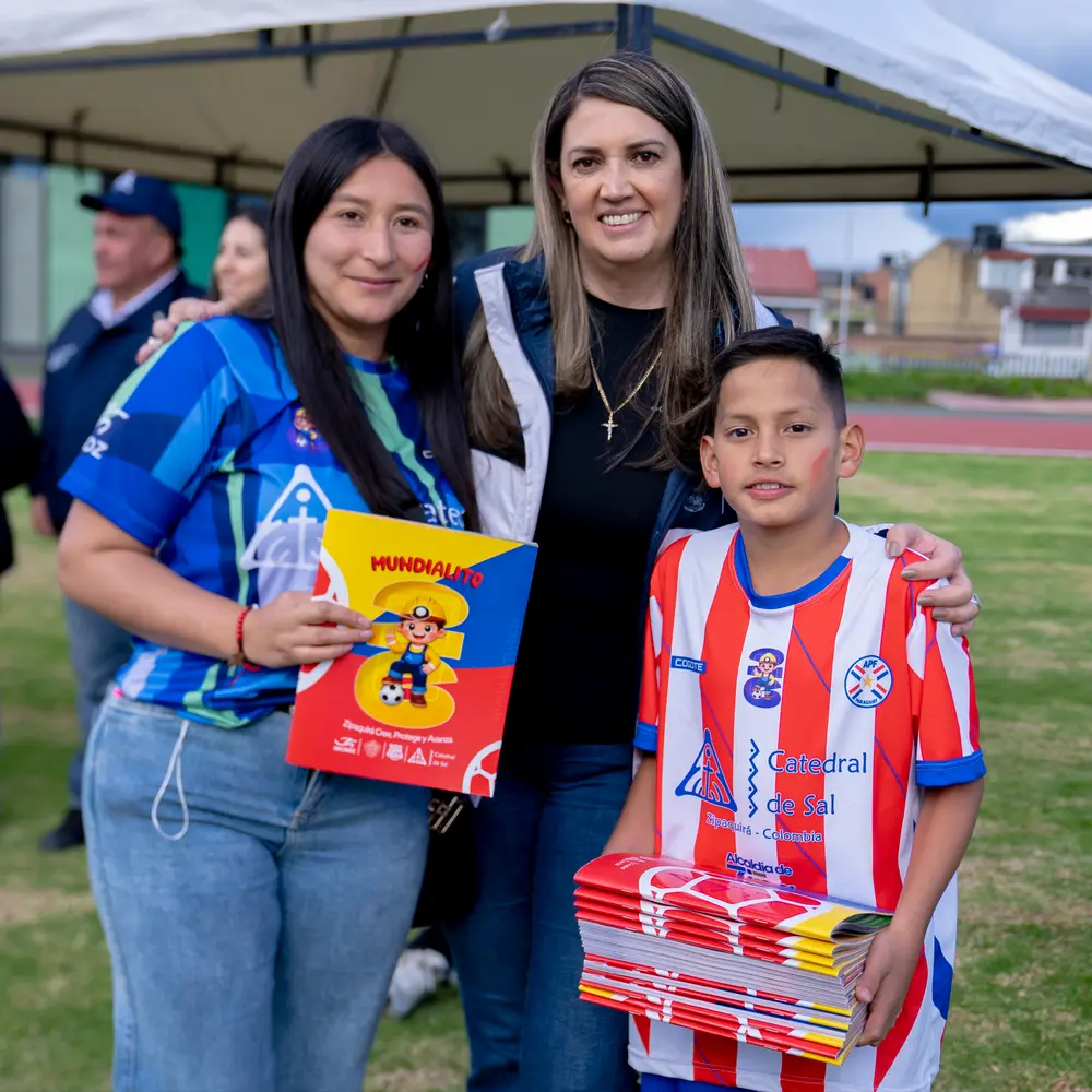 Dos mujeres y un niño futbolista sonríen mientras muestran los álbumes oficiales del torneo Mundialito 2026 patrocinado por la Catedral de Sal de Zipaquirá.