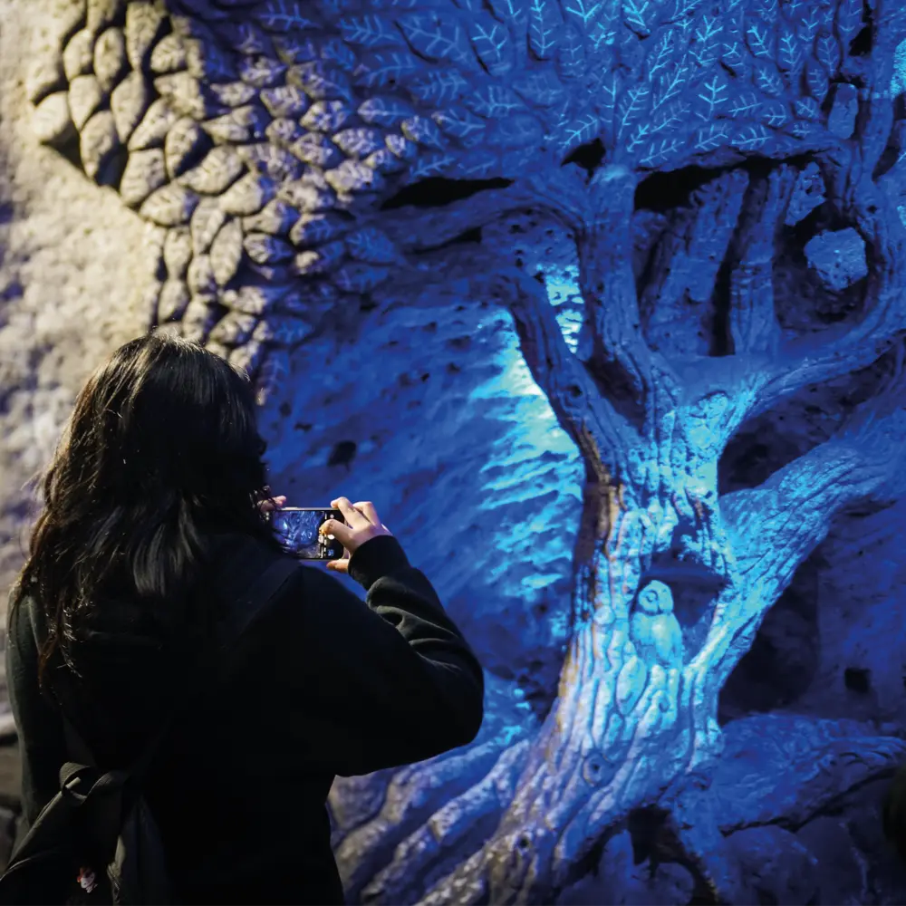 Una mujer toma una foto con su celular a una escultura de un árbol tallado en roca de sal con iluminación azul en la Catedral de Sal de Zipaquirá.