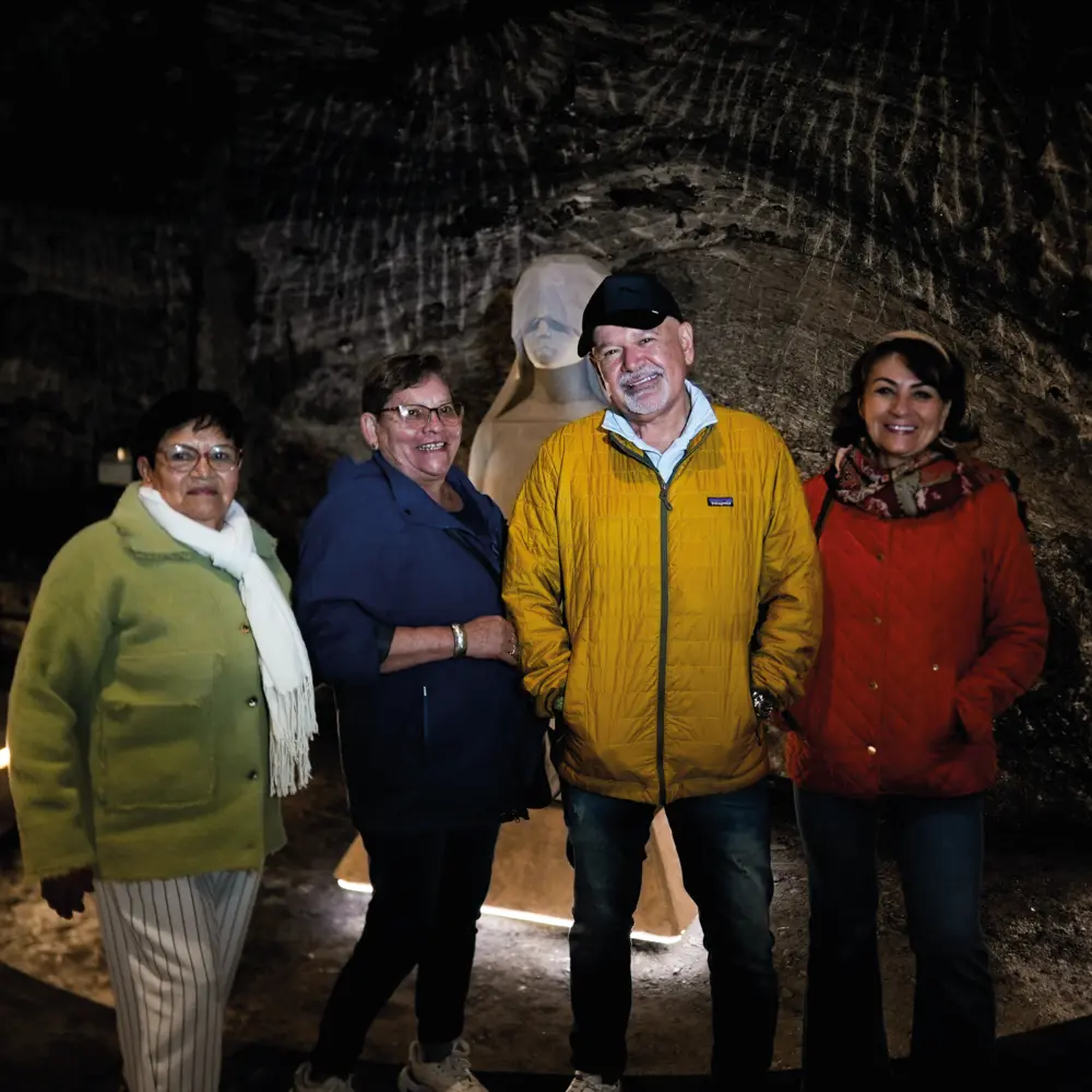 Un grupo de adultos mayores sonríe posando junto a la escultura iluminada de la Virgen María tallada en sal dentro de la Catedral de Sal.