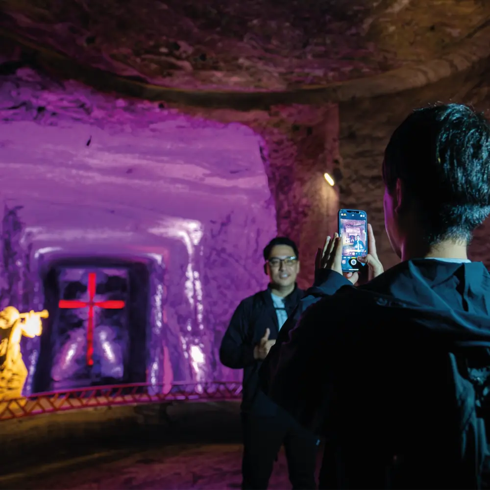 Visitante toma una fotografía con su celular a un turista en un altar iluminado con luces purpuras y una cruz roja al fondo en la Catedral de Sal.