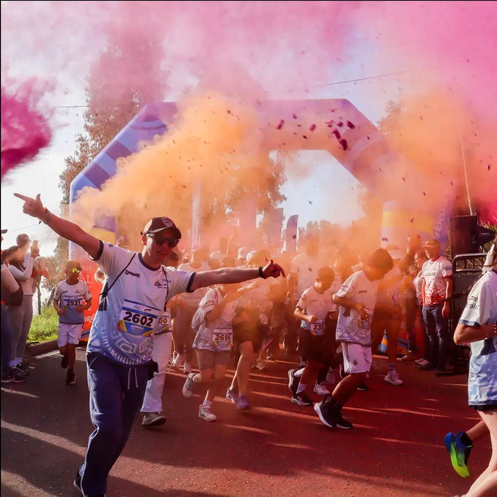 Fotografía de la salida masiva en la carrera Trail Running de Colores en Zipaquirá. Muestra a corredores de todas las edades cruzando el arco inflable en medio de una intensa nube de polvos holi color rosa y naranja