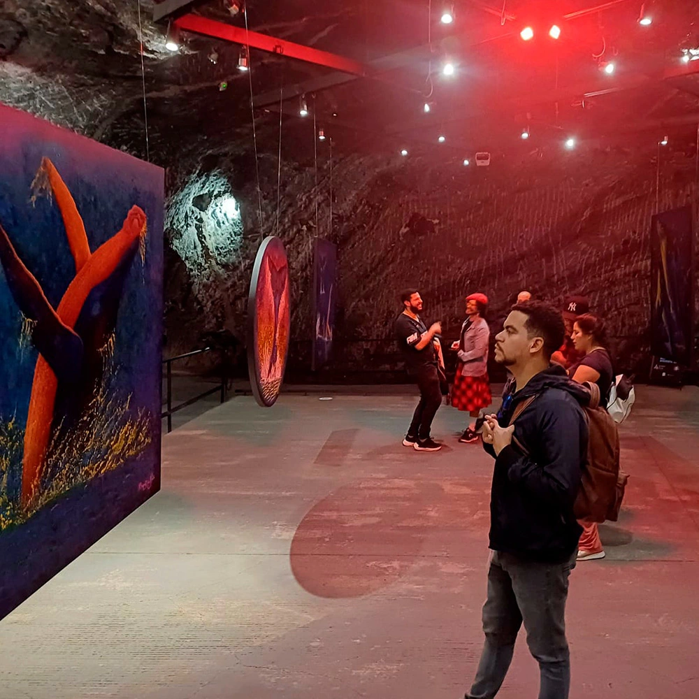 Un joven con mochila observa una pintura detallada de la cola de una ballena en la exposición 'Yubartas Gigantes Gentiles' dentro de la Catedral de Sal de Zipaquirá, con otros visitantes y cuadros flotantes iluminados bajo luces rojas al fondo