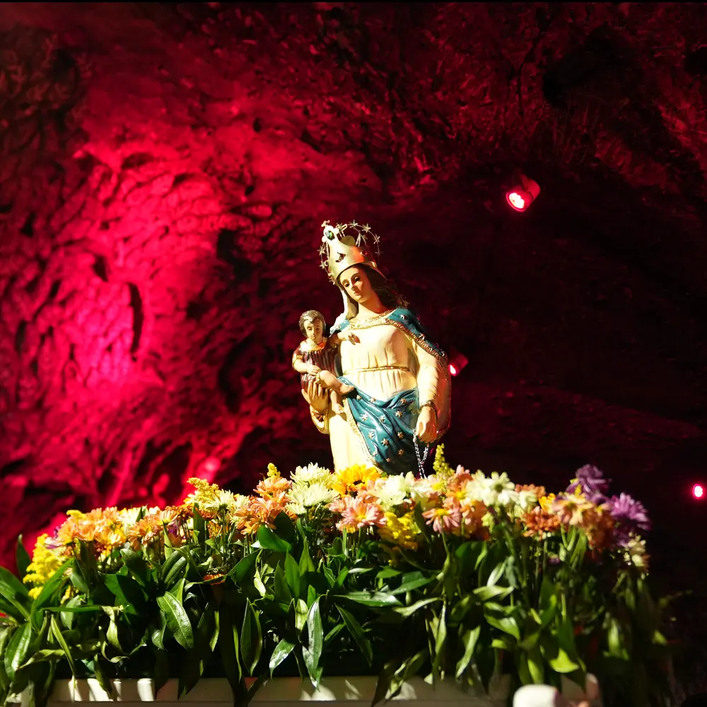 Close-up de la escultura de la Virgen de Guasá con el Niño Jesús sobre un arreglo floral de margaritas, destacando sobre las paredes de roca salina de la Catedral de Sal iluminadas en tono rojo.