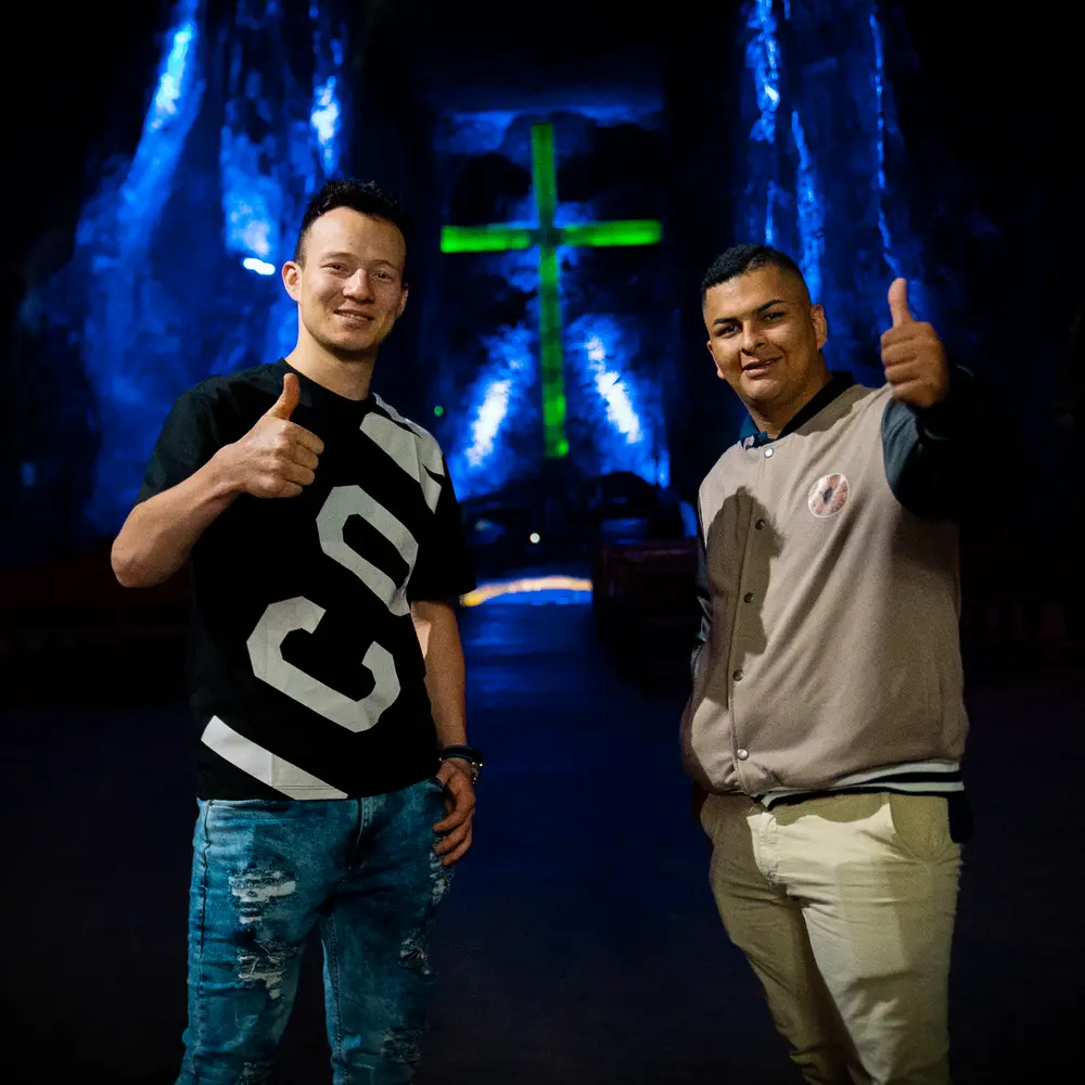 Dos amigos sonriendo y haciendo el gesto de pulgar arriba frente a la gran cruz subterránea iluminada en la Catedral de Sal de Zipaquirá.