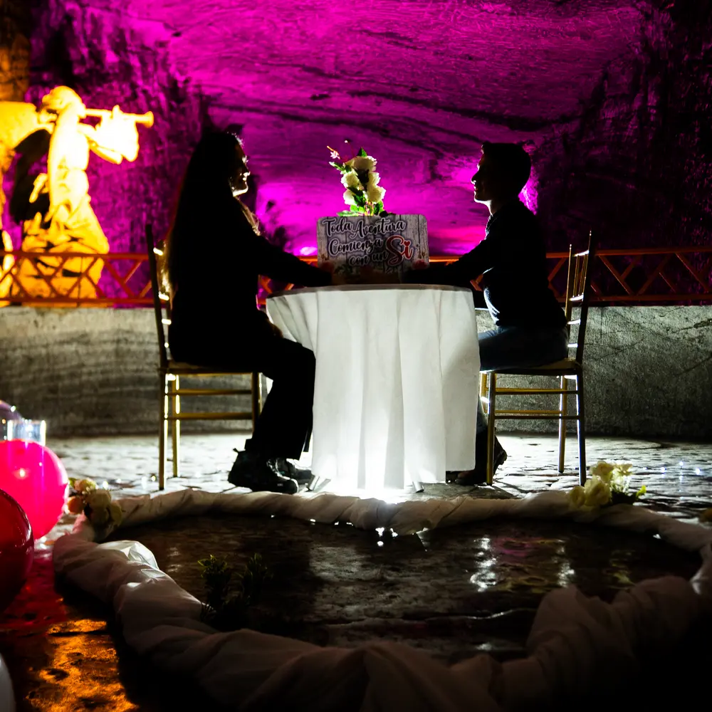 Pareja sentada en una mesa decorada con flores y cartel de propuesta romántica, iluminada bajo tierra con luces magenta durante Amor y Amistad en la Catedral de Sal de Zipaquirá