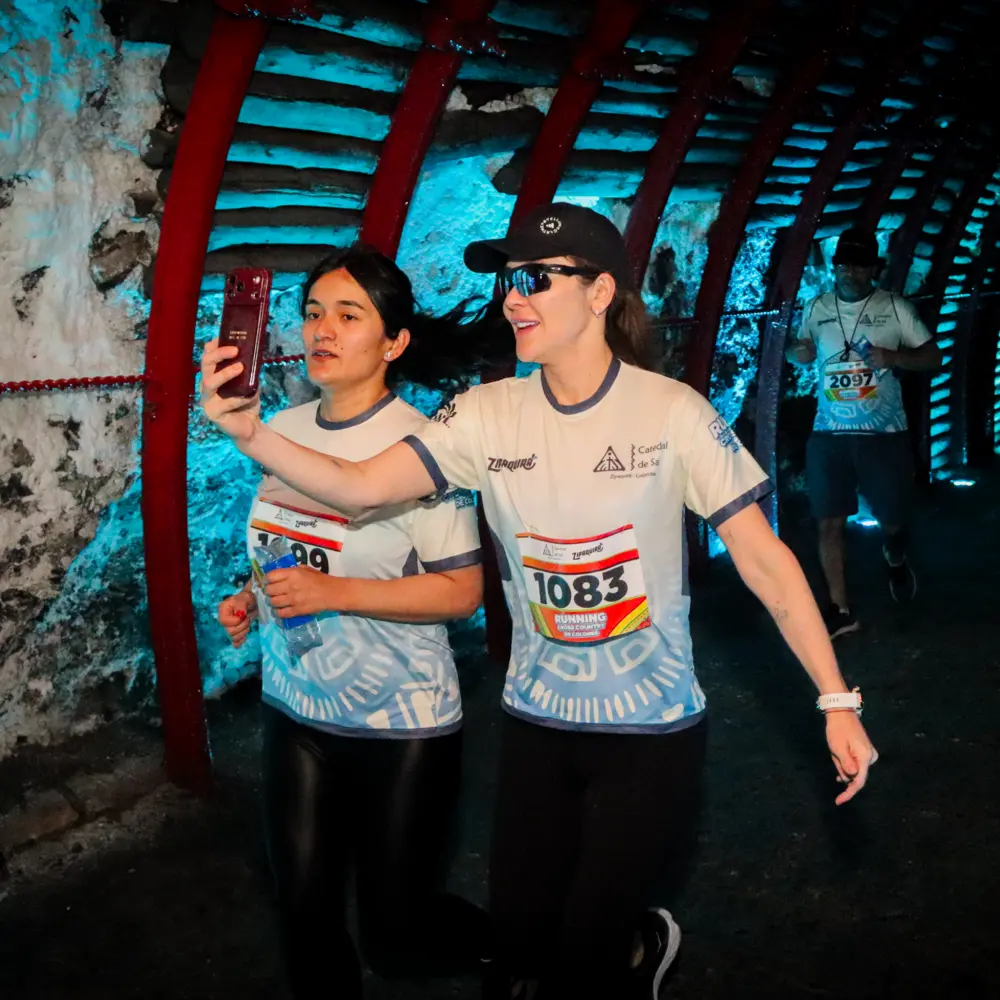 Dos mujeres corredoras tomándose una foto con su celular mientras corren por el túnel subterráneo iluminado de azul en la Catedral de Sal de Zipaquirá durante la carrera Trail Running de Colores 2026