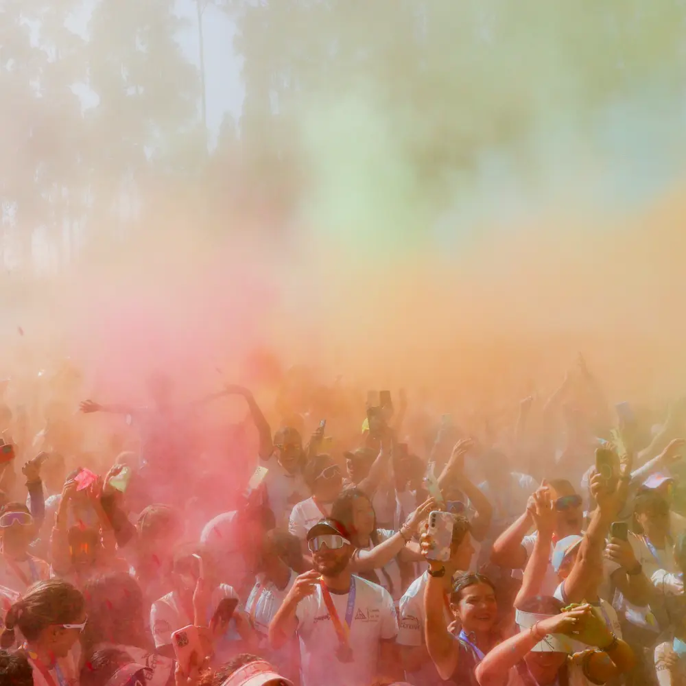 Multitud de personas y corredores reunidos al aire libre envueltos en una gran nube de polvo de colores rosa, naranja y verde durante la celebración del Trail Running de Colores 2026