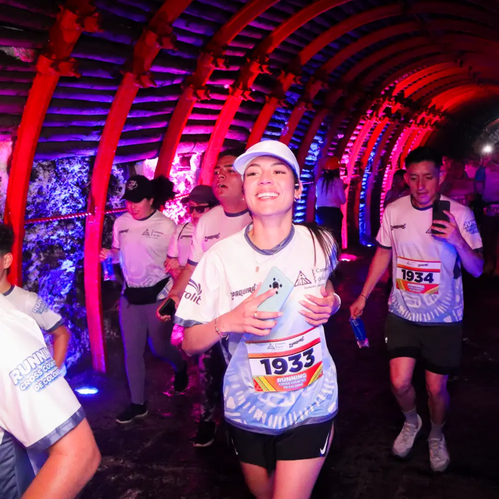 Una mujer sonriente corriendo en primer plano junto a otros participantes dentro del socavón subterráneo iluminado con luces moradas y rojas durante la carrera Trail Running de Colores 2026 en la Catedral de Sal.