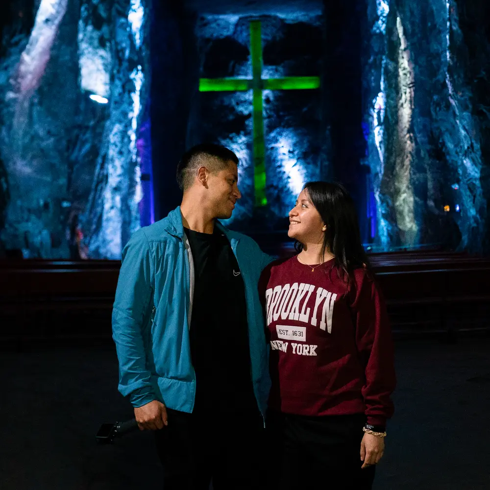 Pareja sonriendo y mirándose fijamente frente a la imponente cruz subterránea iluminada en verde dentro de la Catedral de Sal de Zipaquirá