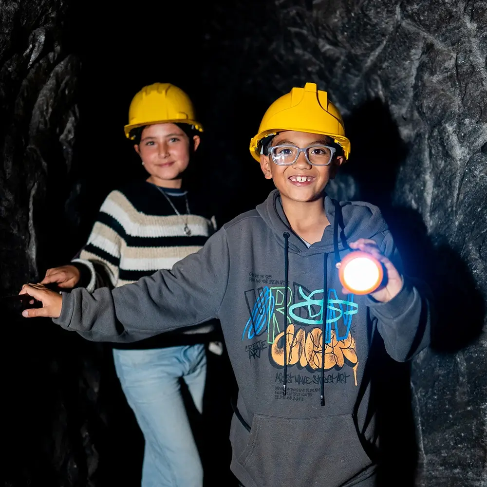 Niño sonriente con gafas y casco amarillo de minero apuntando una linterna encendida mientras explora un túnel subterráneo en la Catedral de Sal de Zipaquirá junto a una niña