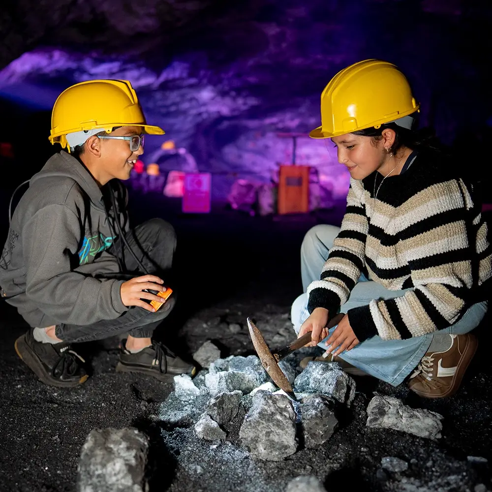 Dos niños sonrientes vistiendo cascos amarillos de minero participando en la actividad de la Ruta del Minero picando rocas de sal en la Catedral de Sal de Zipaquirá