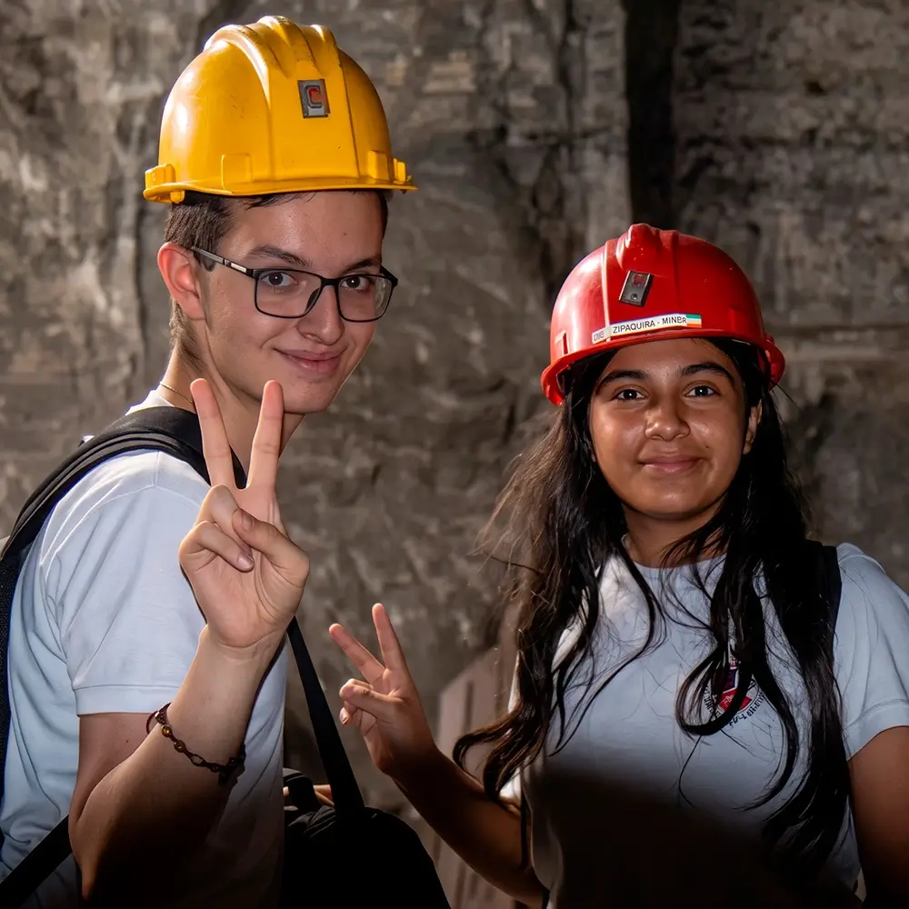 Dos jóvenes sonrientes, un chico con casco amarillo haciendo el signo de la paz y una chica con casco rojo, posando frente a la pared de roca en el interior de la Catedral de Sal de Zipaquirá durante la semana de receso.Dos jóvenes sonrientes, un chico con casco amarillo haciendo el signo de la paz y una chica con casco rojo, posando frente a la pared de roca en el interior de la Catedral de Sal de Zipaquirá durante la semana de receso.
