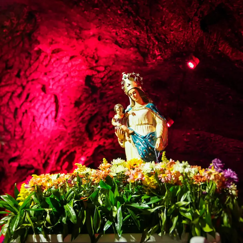 Plano medio de la Virgen del Rosario de Guasá con el Niño Jesús sobre un arreglo floral festivo, resaltada con iluminación roja en las paredes de sal de la Catedral de Sal de Zipaquirá